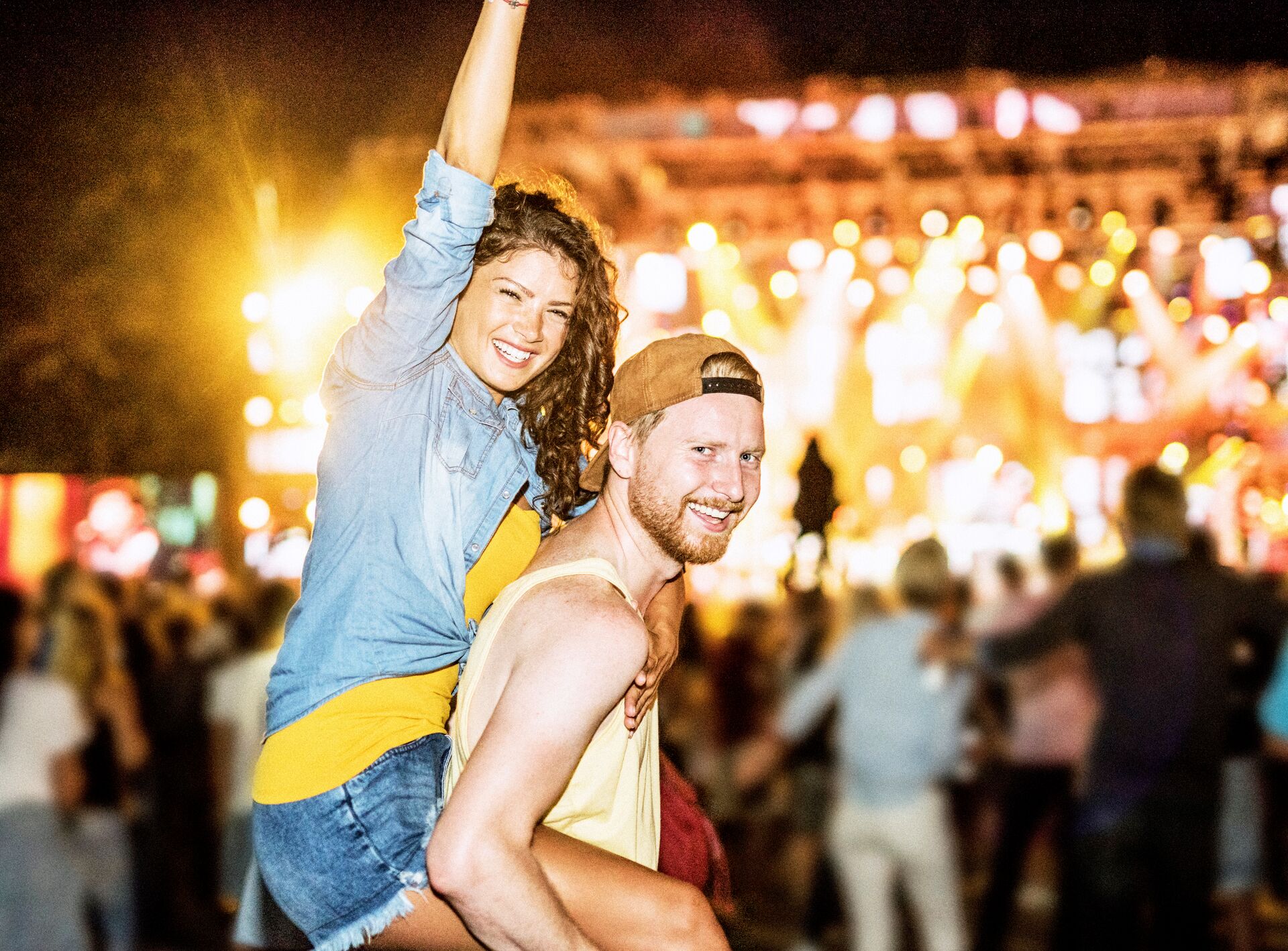 Large_MS PPT_Web-A happy young man with a hat giving his female friend a piggyback ride while smiling at the camera at a music festival at night.jpg