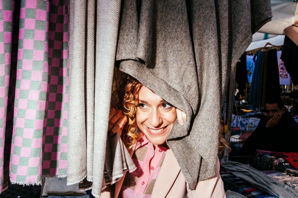 Large_Web and Screen-A woman smiles as she peeks out from behind woven scarves at a market stand.jpg