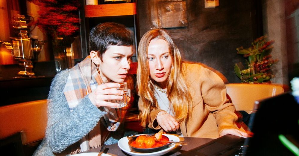 Two female friends sit at a restaurant table with food and look at a laptop.jpg