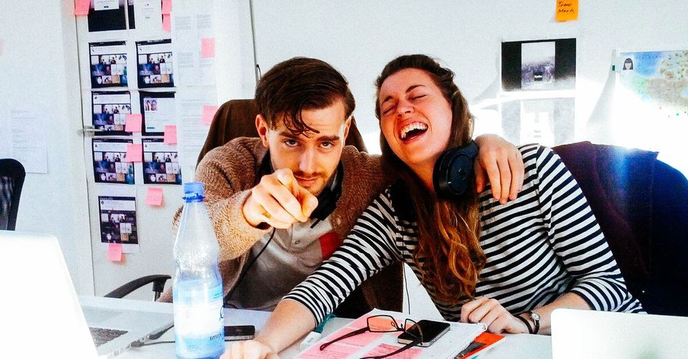 Young man and woman laughing and pointing at the camera while sitting at a desk with laptops.jpg